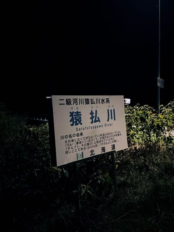 夜の草むらに立つ白い河川標識。二級河川猿払川水系、猿払川、Sarufutsugawa River と書かれ、その下に川の名の由来が記されている。