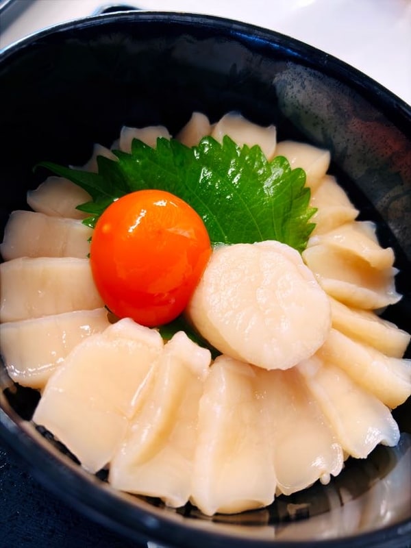 黒い丼に帆立の貝柱が放射状に並び、中央に卵黄と青い葉が置かれている。