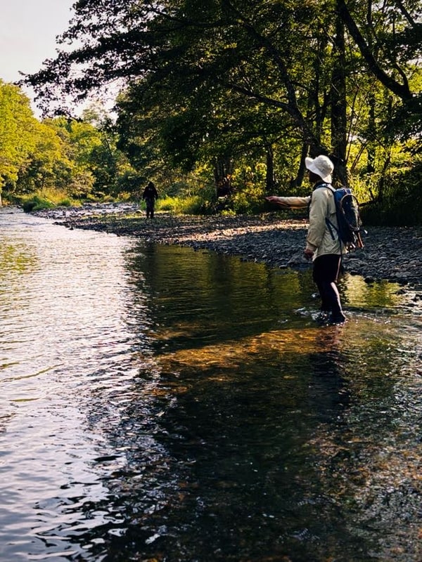 森に挟まれた浅い川に、胴長の人物が二人立っている。手前の一人は背を向け、奥の一人は遠い。顔は写っていない。