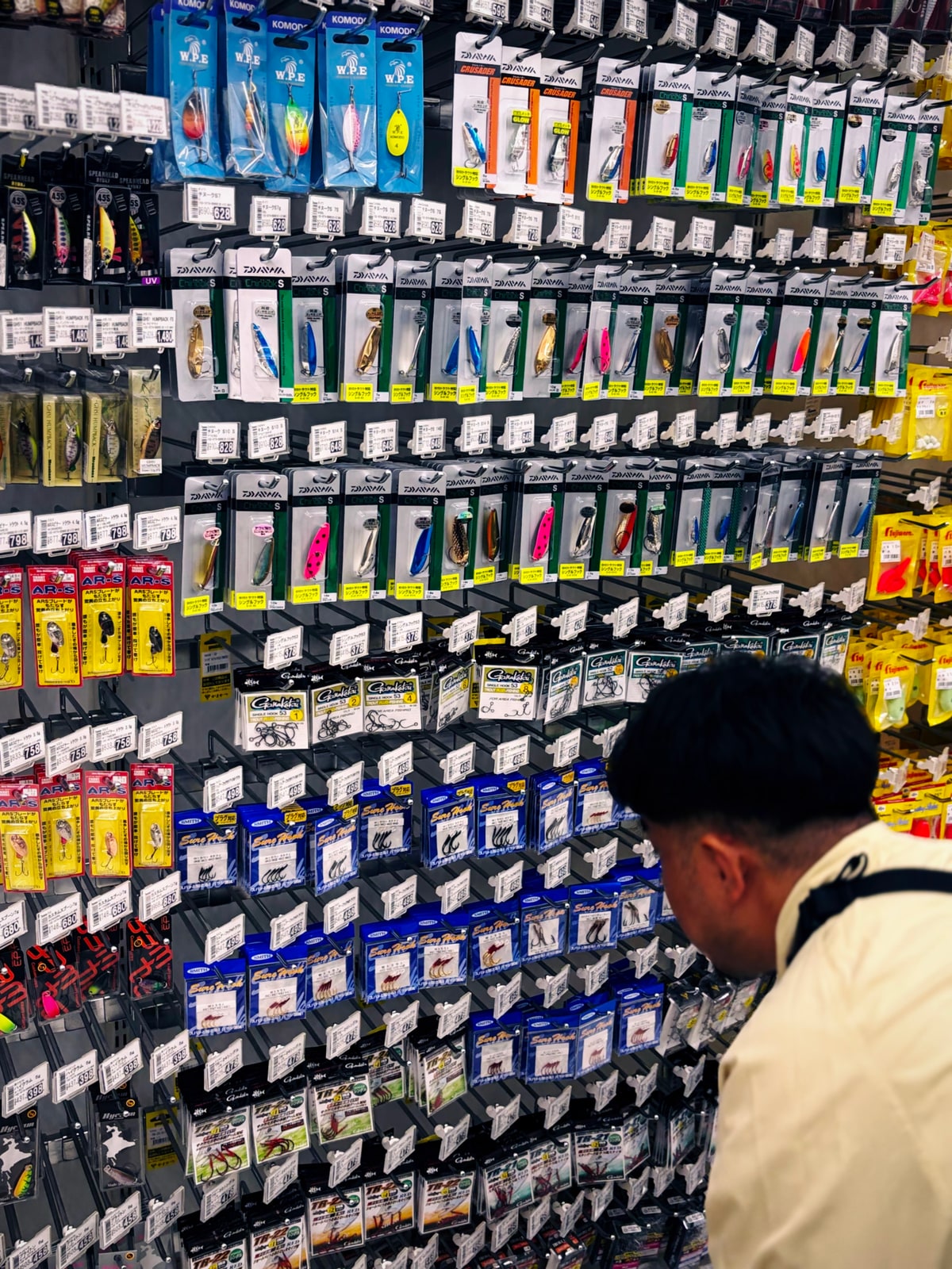 別の釣具店の壁一面のルアー。手前の人物が背を向けて見上げている。顔は写っていない。