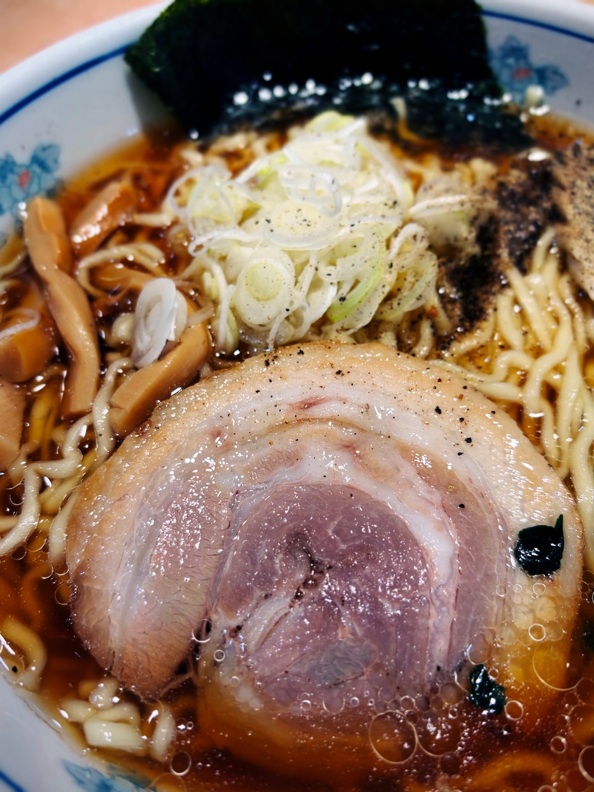 青い花柄の丼に醤油色の澄んだ汁のラーメン。巻いた叉焼が一枚、白髪に切った葱、メンマ、海苔、黒胡椒。