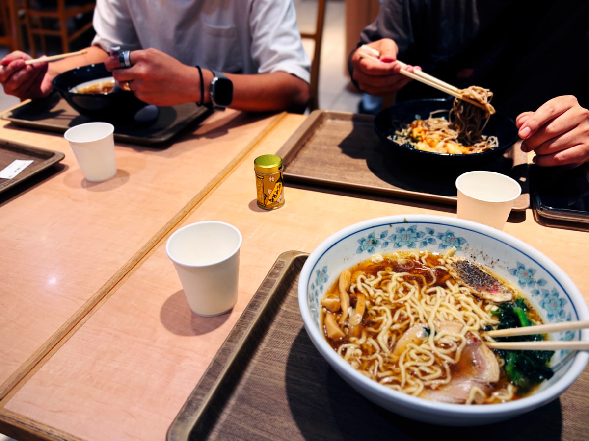 フードコートの木の卓。手前の盆にラーメンの丼、向かいでは二人が黒い丼から麺を取っている。卓の中ほどに紙コップと小さな薬味の瓶。