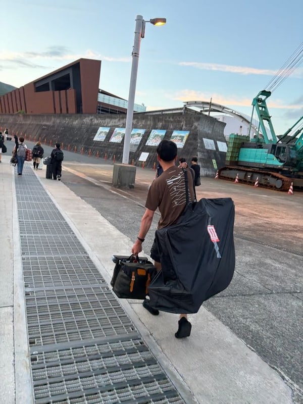 明け方の港の岸壁を、大きな黒い荷と道具箱を提げた人が後ろ姿で歩いている。奥に緑色のクレーンとターミナルの建物。