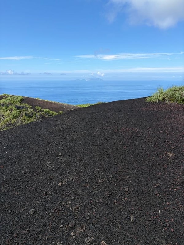 手前に黒いスコリアの緩い斜面が広がり、その向こうの青い海に、平たい頭に雲を載せた島影が一つ浮かんでいる。