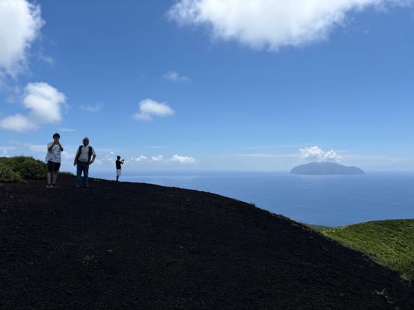 黒いスコリアの尾根の上に三人が離れて立ち、海の方を向いている。右手の水平線に島影が一つ。