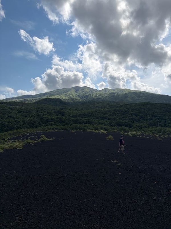 一面の黒いスコリアの野原を人が一人横切っている。奥に緑の斜面が重なり、その上に雄山と大きな積雲。
