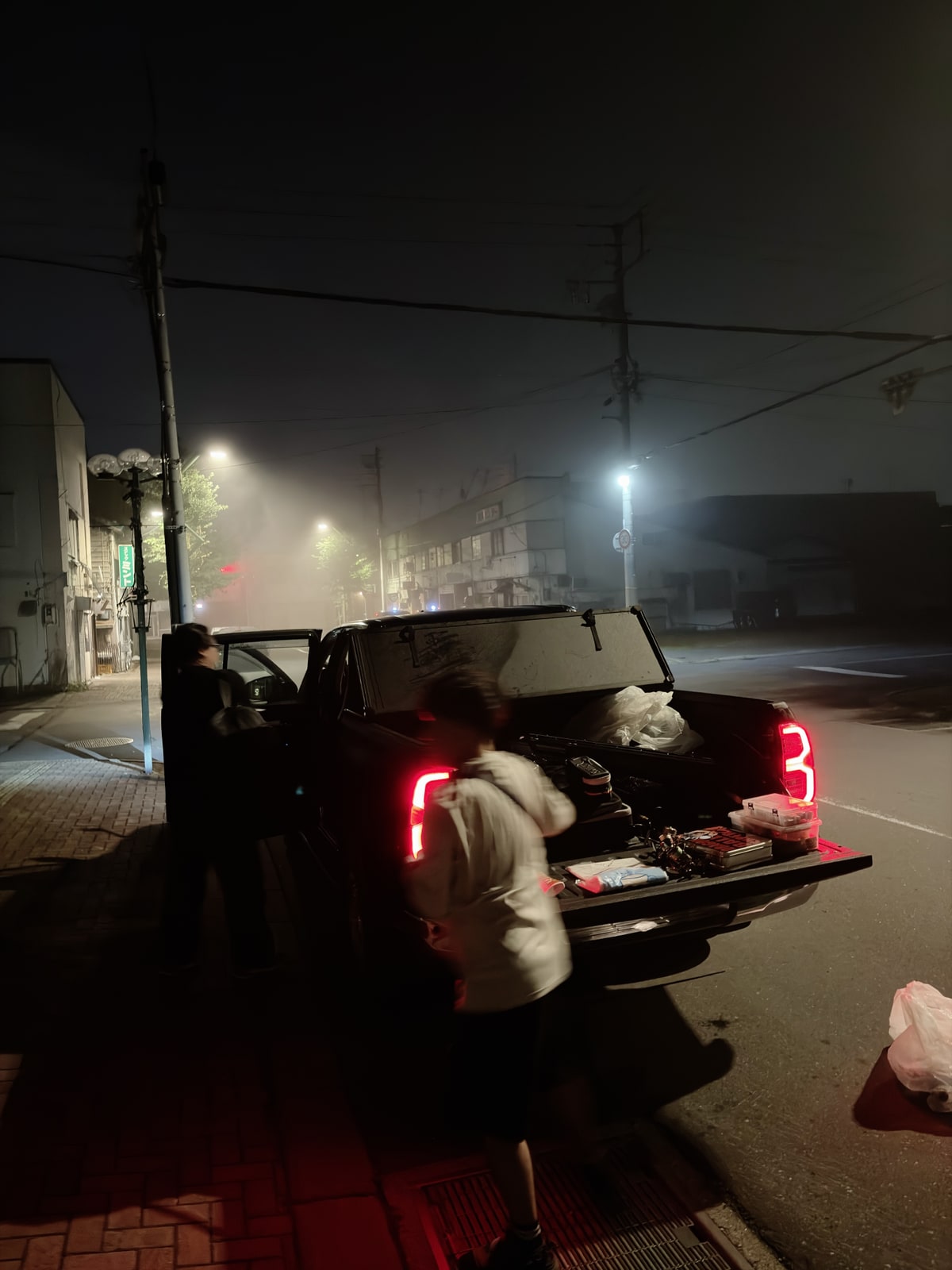深夜の街灯の下、荷台の蓋を開けた黒いピックアップ。尾灯が赤く点き、濡れた舗装に赤が伸びている。荷台で人が荷を分けている。顔は写っていない。
