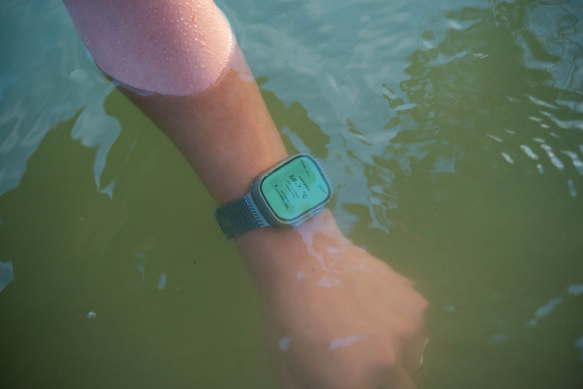 緑がかった湖の水に前腕が沈み、腕時計の画面が水面の下で緑に光っている。肘のあたりで水の膜が切れている。