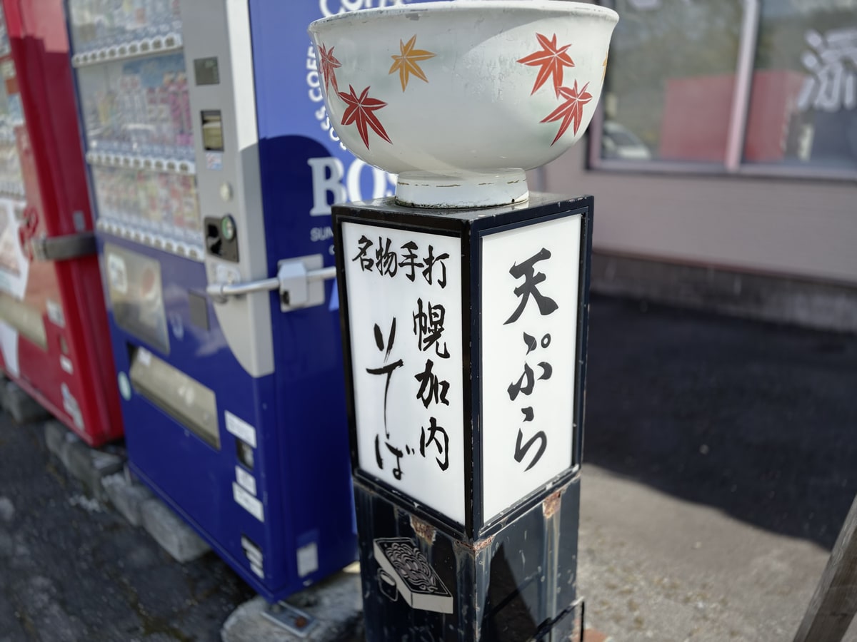 道端の自動販売機の横に立つ行灯の看板。手打ちのそばと天ぷらの文字が入っている。