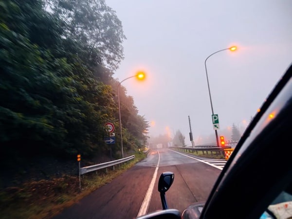 雨のフロントガラス越しに見る朝の高速道路。路肩の木立が灰色に沈み、前を行く車の尾灯が滲んでいる。