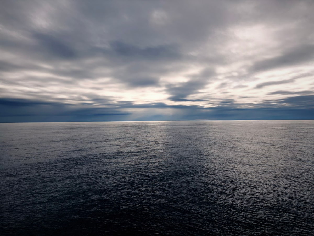 厚い雲の帯の切れ目から光が差し、暗い海の一部だけが帯のように白く光っている。