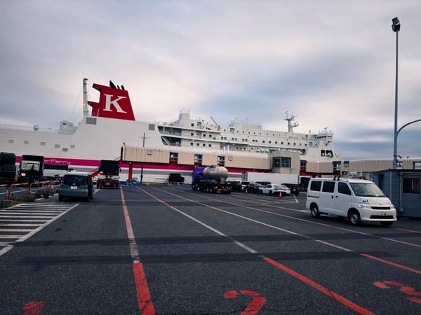 曇りの港の広い舗装。奥に白と赤のフェリーが停まり、手前に車が数台。